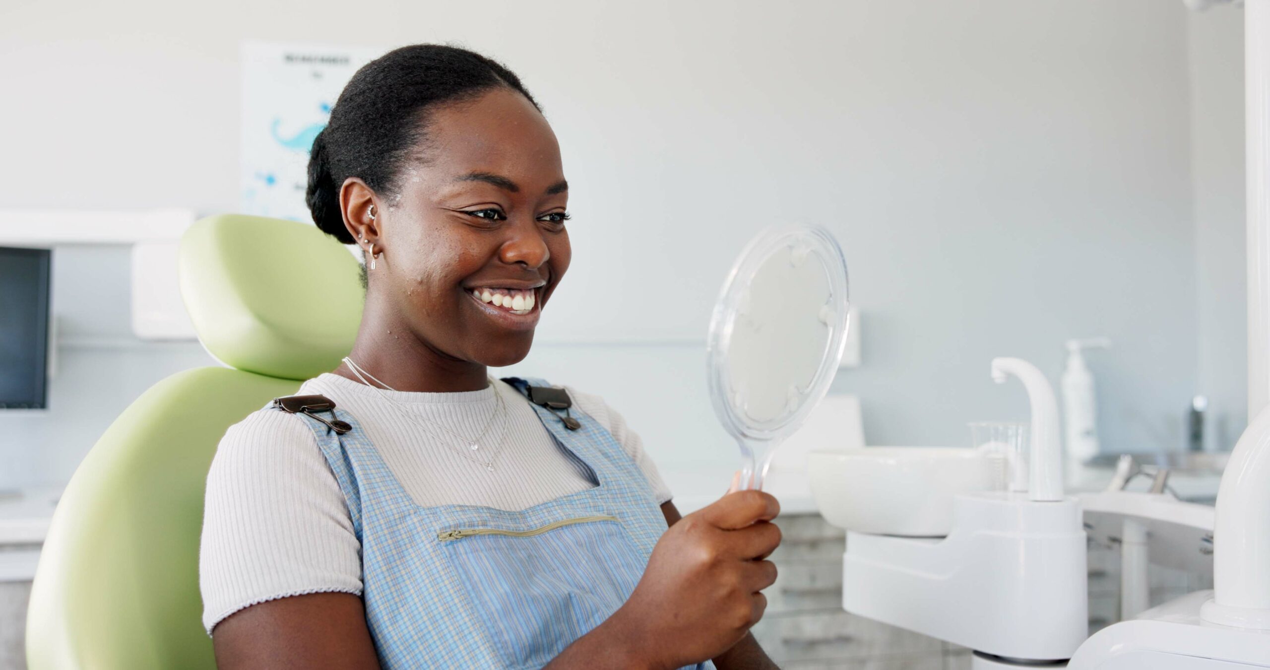 happy young woman sitting in a dental hair, smiling into hand mirror